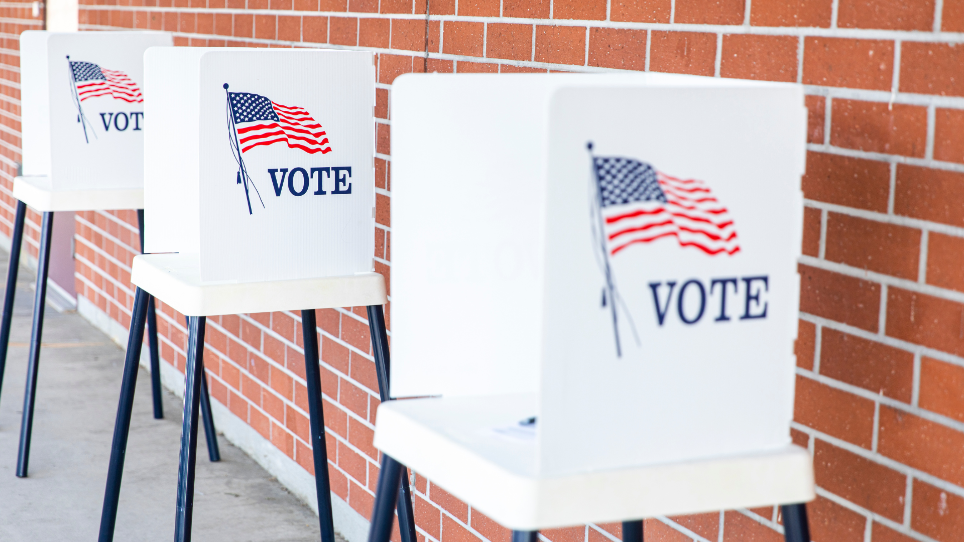 Three standalone voting booths in a line against a brick wall.
